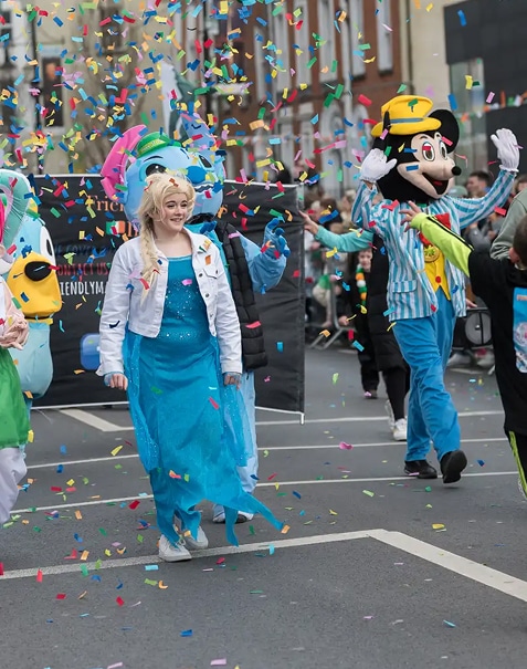 A person dressed as Elsa from Frozen and others in cartoon character costumes walk in a street parade, surrounded by colourful confetti falling in the air.