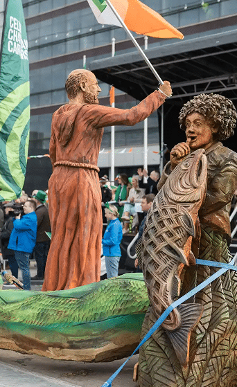A parade float features two large wooden sculptures: a man in a robe holding an Irish flag and a woman with curly hair holding a carved fish. People and photographers watch in the background.