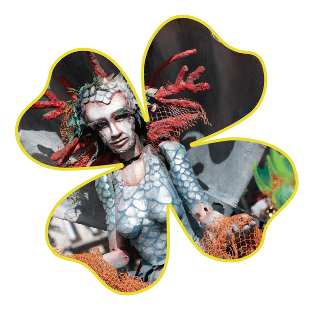 A person in a detailed sea-inspired costume with scales, coral-like headpiece, and orange netting is seen through a clover-shaped frame with a yellow outline.