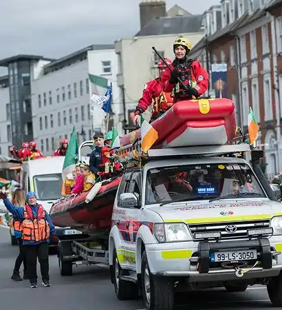 A parade scene at the St Patrick’s Day Festival Waterford shows people in life jackets and helmets riding a rescue boat mounted on a lorry, with Irish flags on display. Buildings line the street as spectators watch the festive procession.