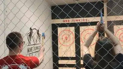 Two people stand behind a chain-link fence, preparing to throw axes at wooden targets marked with red bullseyes in "Axe Junkies," a perfect spot to visit after you explore Waterford St Patrick’s Day Festival.