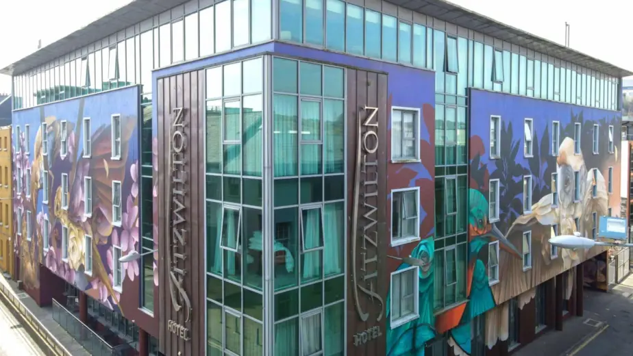 A modern hotel building with large glass windows and corner signage reading "FITZWILTON HOTEL"; its exterior features colourful mural art—an ideal base to explore Waterford St Patrick’s Day Festival.