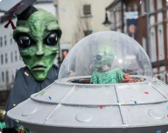 Two people in alien costumes join the St Patrick’s Day Festival Waterford parade; one sports a large green alien mask with a graduation cap, whilst the other, dressed as a small alien, sits inside a UFO prop as confetti falls around them.