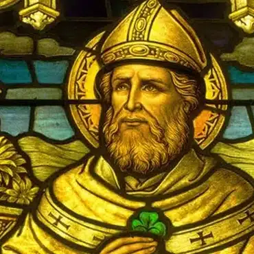 A stained glass image of a bearded man in a gold bishop’s hat and robe, with a halo, holding a green shamrock—reflecting the spirit of Demilitarising Saint Patrick’s Day 1921.