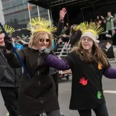 Two people in festive outfits with yellow headpieces and colourful handprints on their shirts smile and dance together during an outdoor parade, celebrating Demilitarising Saint Patrick’s Day 1921 among other joyful participants and onlookers.
