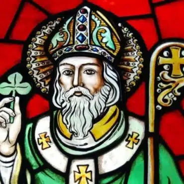 Stained glass artwork of a bearded saint, evoking Demilitarising Saint Patrick’s Day 1921, wearing a bishop’s hat and green robes, holding a shamrock and gold staff against a vivid red background.