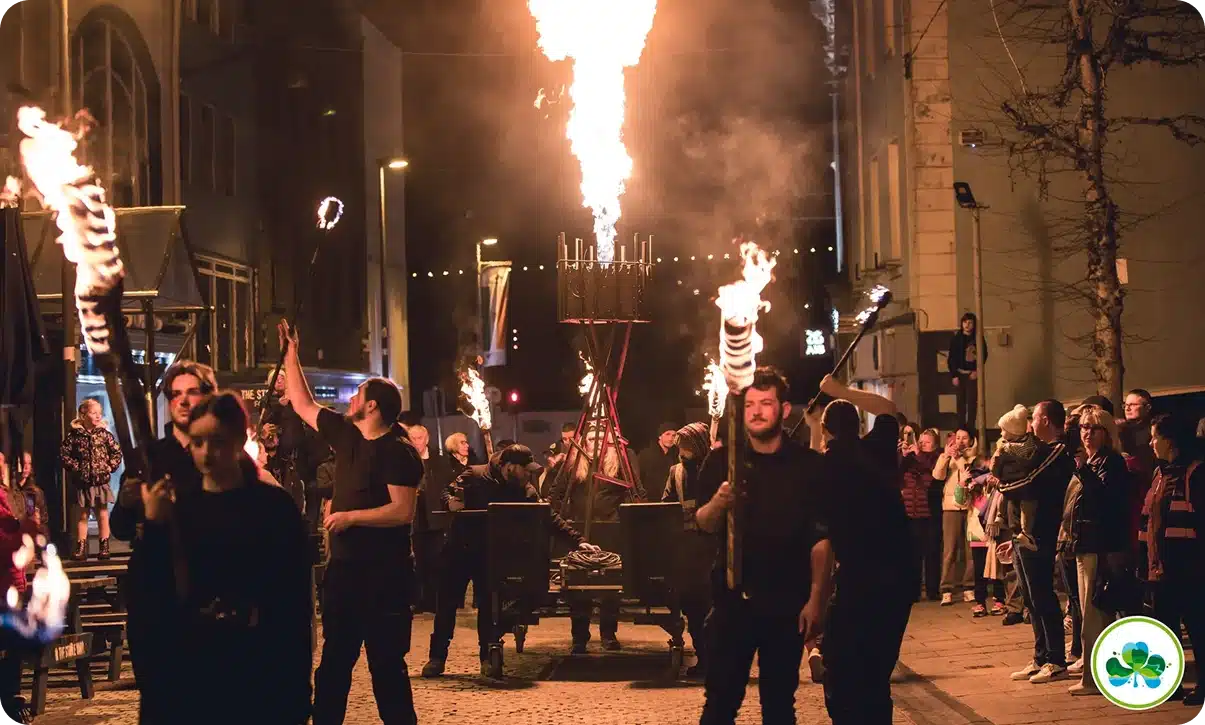 A night-time street scene shows people in black holding torches, with a flaming structure in the centre. Spectators line the street, watching the fire display. Buildings and lights are visible in the background.