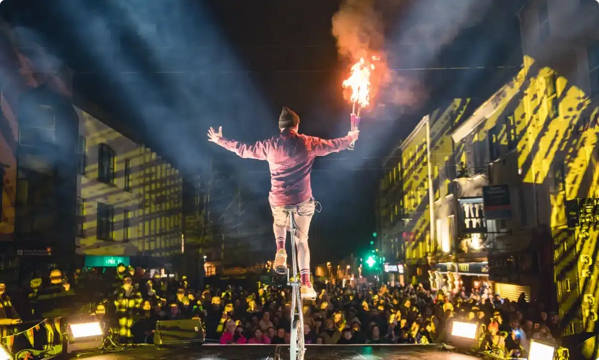 A performer balances on a tall unicycle, arms outstretched, holding a flaming torch before a large crowd at the St Patrick’s Day Festival Waterford, with bright spotlights and city buildings illuminated in the background.