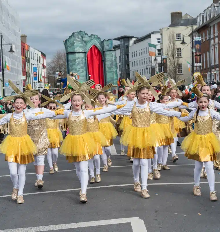 A group of girls in gold and yellow costumes dance in formation during the St Patrick’s Day Festival Waterford parade, with spectators, festive floats, and sponsors’ banners in the background.