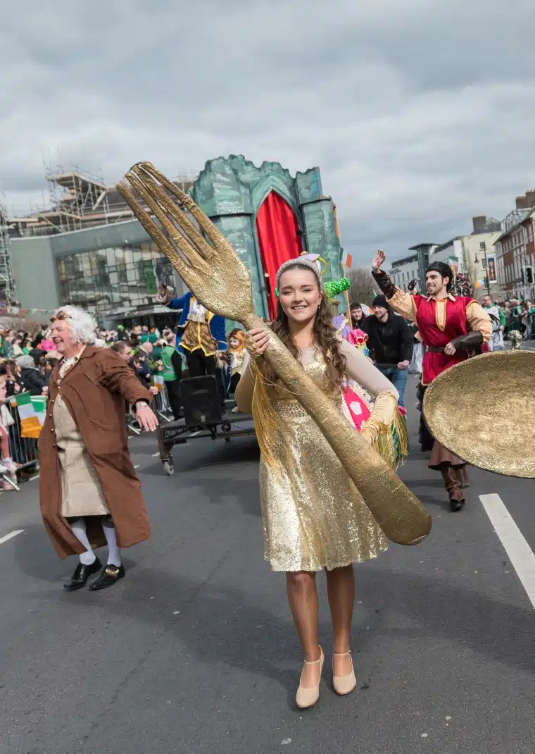 A smiling woman in a gold dress holds an oversized gold fork, leading the St Patrick’s Day Festival Waterford parade with costumed participants, including people in historical outfits, on a city street under a cloudy sky.