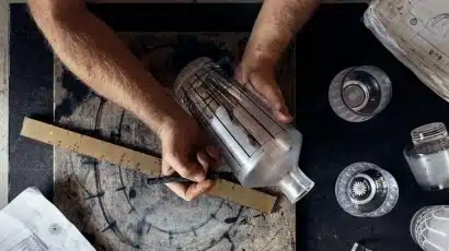A person uses a ruler and pen to mark precise lines on a glass bottle. The workspace, ideal for those who explore Waterford St Patrick’s Day Festival, includes a circular diagram, technical drawings, and other glass objects.