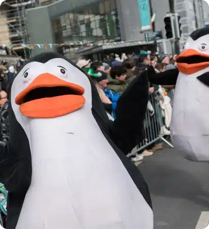 Two people in large penguin costumes walk in the St Patrick’s Day Festival Waterford parade, surrounded by crowds behind metal barriers. The busy, decorated street is filled with onlookers enjoying the lively celebration.