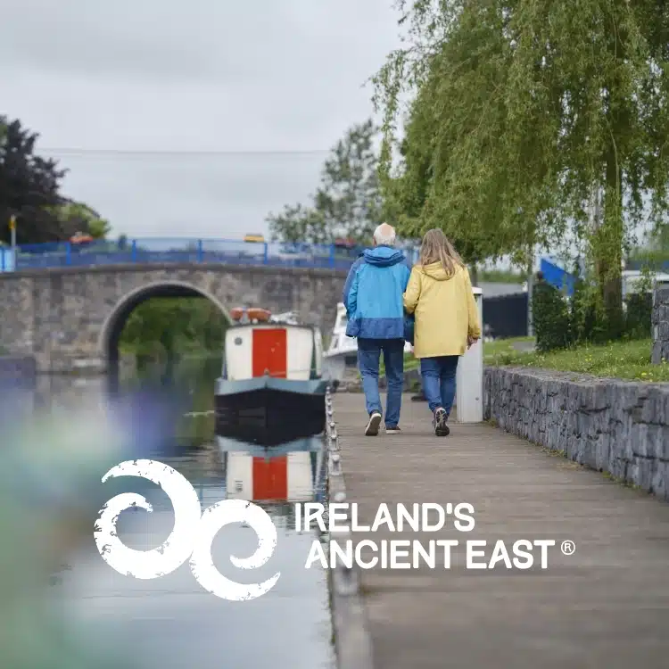 An older couple walk hand-in-hand along a canal towpath beside a boat, with an arched stone bridge and greenery in the background. The text reads "Ireland’s Ancient East—Explore Waterford St Patrick’s Day Festival.