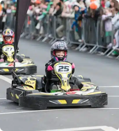 A young child wearing a helmet drives a go-kart labelled with the number 25 in a race at the St Patrick’s Day Festival Waterford, with another child in kart number 27 behind and crowds of spectators watching from behind barriers.