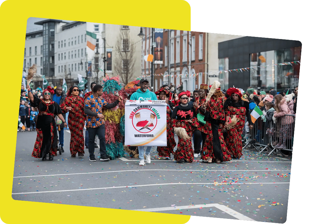 A group of people in colourful traditional clothing march in the St Patrick’s Day Festival Waterford parade, holding a banner that reads "Nigerian Community Waterford" as spectators watch from behind barriers. Confetti is scattered on the street.