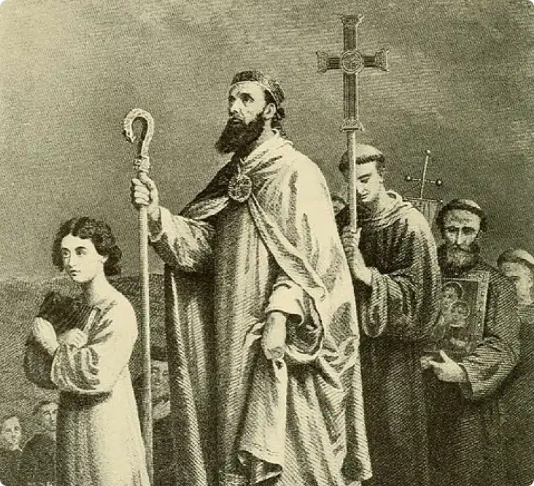 A group of people in mediaeval attire walk forward; a bearded man in the centre wears a robe and carries a staff, followed by others holding a cross and religious icon, evoking the solemn history of St Patrick Ireland.
