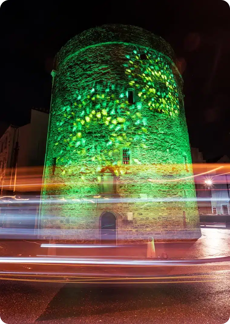 A round stone tower is illuminated with green and yellow lights at night during the St Patrick’s Day Festival Waterford, with streaks of car lights passing by in front, creating a dynamic effect.