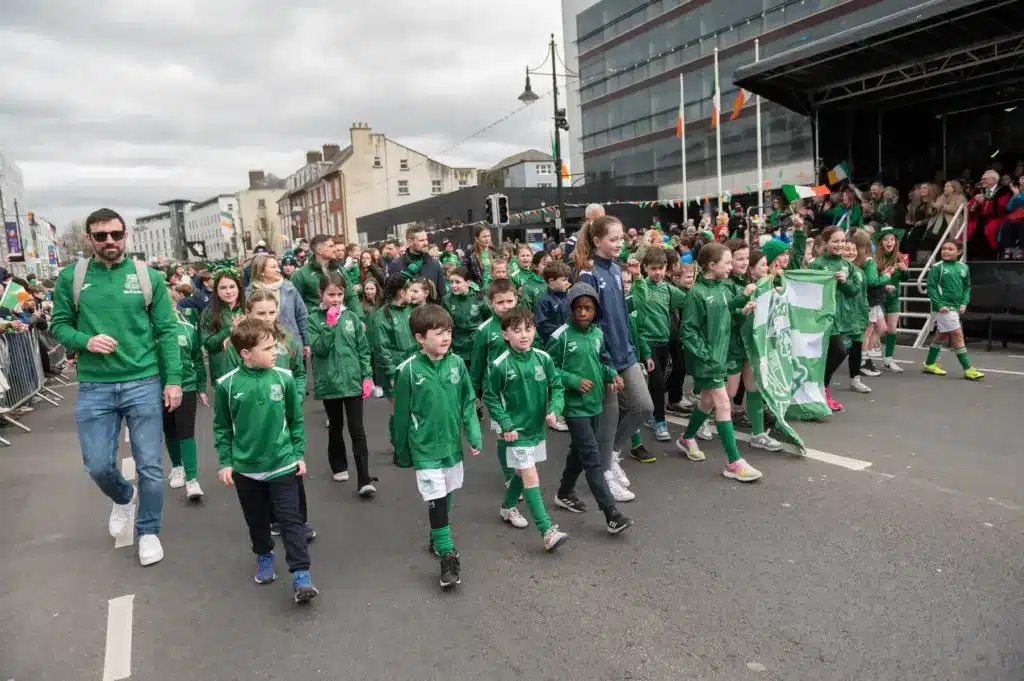 A large group of children and adults in matching green sports jackets walk together in a street parade during the St Patrick’s Day Festival, cheered on by a crowd. Flags and festive decorations can be seen in the background.
