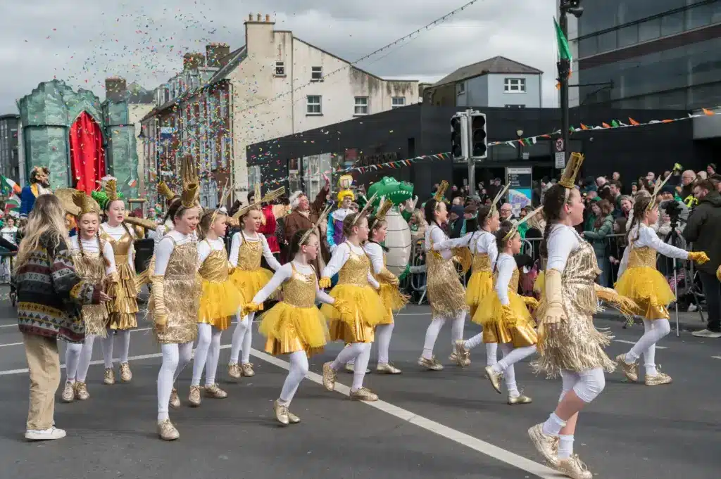 A group of children in yellow and gold costumes dance in a parade during the St Patrick’s Day Festival, as a crowd watches and colourful confetti falls along a lively Waterford city street.