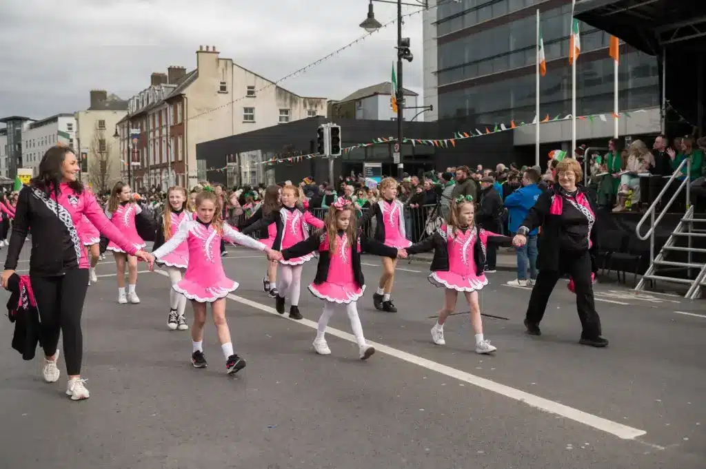 A group of young girls in matching pink and white Irish dance costumes hold hands and walk in the St Patrick’s Day Festival parade, accompanied by adults. Spectators watch from the sidelines, with Irish flags waving near the Waterford Gallery.