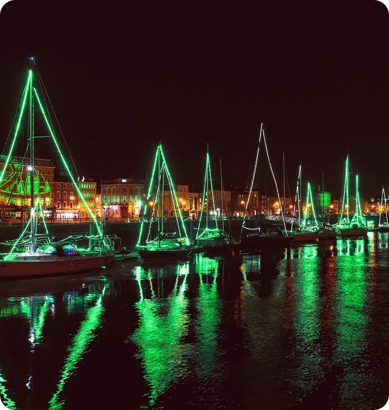 Sailing boats decorated with bright green lights for the St Patrick’s Day Festival Waterford are moored along a waterfront at night, their reflections vivid on the calm water. Buildings lit with warm yellow and orange tones line the background.