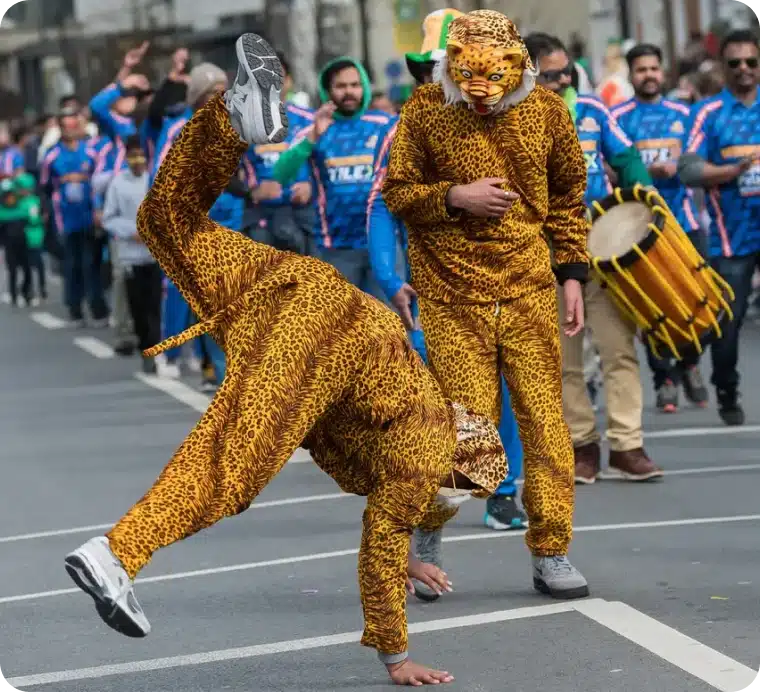 Two people in leopard costumes, one doing a handstand and the other standing with a drum, participate in a lively street parade with a crowd of onlookers in blue shirts in the background.