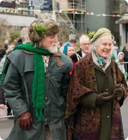 An older man and woman, both smiling, walk outdoors at a parade. The man wears a green scarf and furry hat with greenery; the woman wears a green hat and patterned shawl. People and festive decorations are visible in the background.