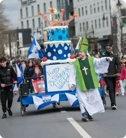 A man dressed as a bishop walks in a parade, leading a float shaped like a giant blue and white birthday cake with “Happy Birthday Ireland” written on it, surrounded by people and flags on a city street.