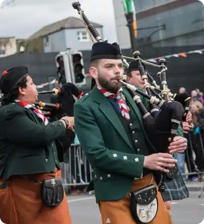 Two musicians wearing green jackets, brown kilts, and hats play bagpipes whilst marching in a parade. People watch from behind barriers and festive bunting hangs in the background.