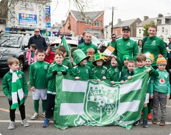 A group of children and adults in green sportswear hold a "Villa F.C." banner and Irish flags at an outdoor event, likely a parade, with parked cars and colourful buildings in the background.