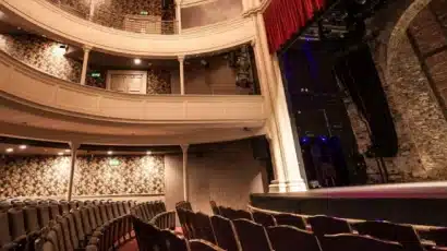 An empty theatre with rows of seats, a curved balcony, ornate wallpaper, and a stage with a red curtain partially open revealing brick walls—perfect for those looking to explore Waterford St Patrick’s Day Festival venues in classic style.