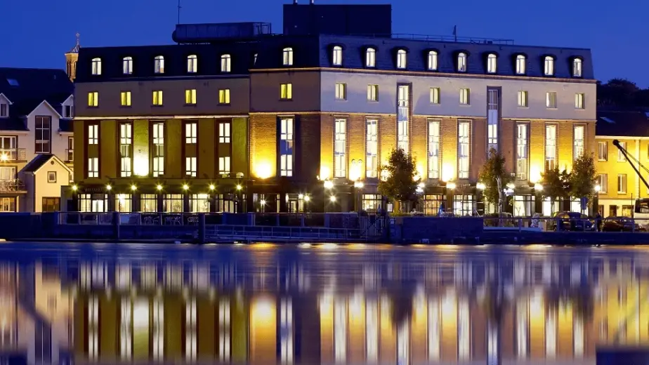 A brightly lit building with many windows is reflected in calm water at night, creating a mirror-like effect. The sky is dark blue, and nearby buildings are also visible—perfect scenery to explore during the Waterford St Patrick’s Day Festival.