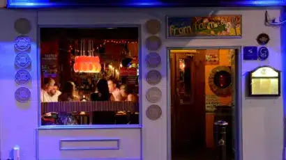 A warmly lit restaurant with people dining inside is seen through a large window at night. The exterior features blue lighting, a sign reading "Bodéga," and a colourful "from farm to fork" sign—perfect for those exploring Waterford St Patrick’s Day Festival.
