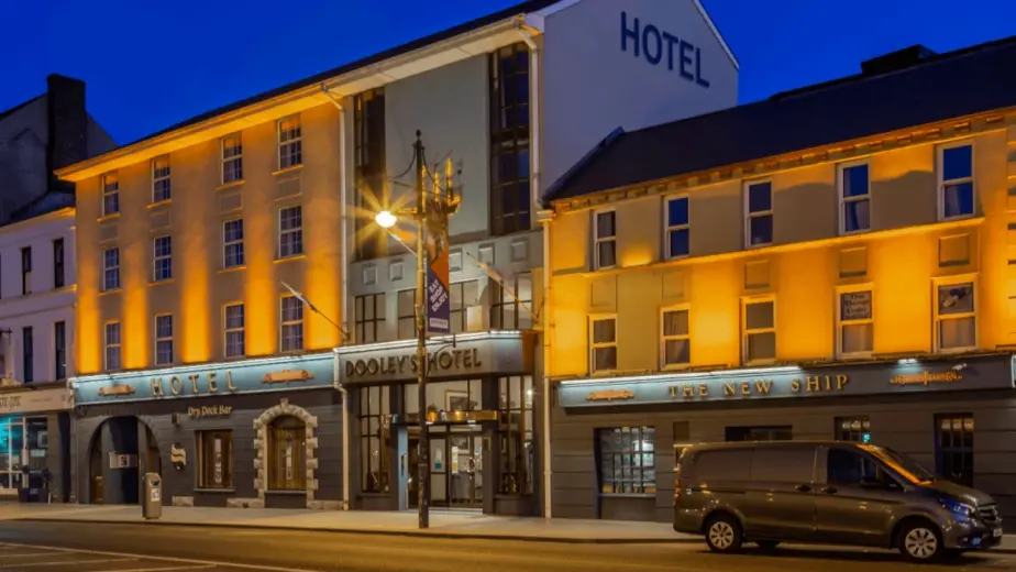 A warmly lit hotel building with a sign reading "Dooley's Hotel" on a city street at dusk welcomes guests to explore Waterford St Patrick’s Day Festival; a van is parked in front, and the sky is a deep blue.