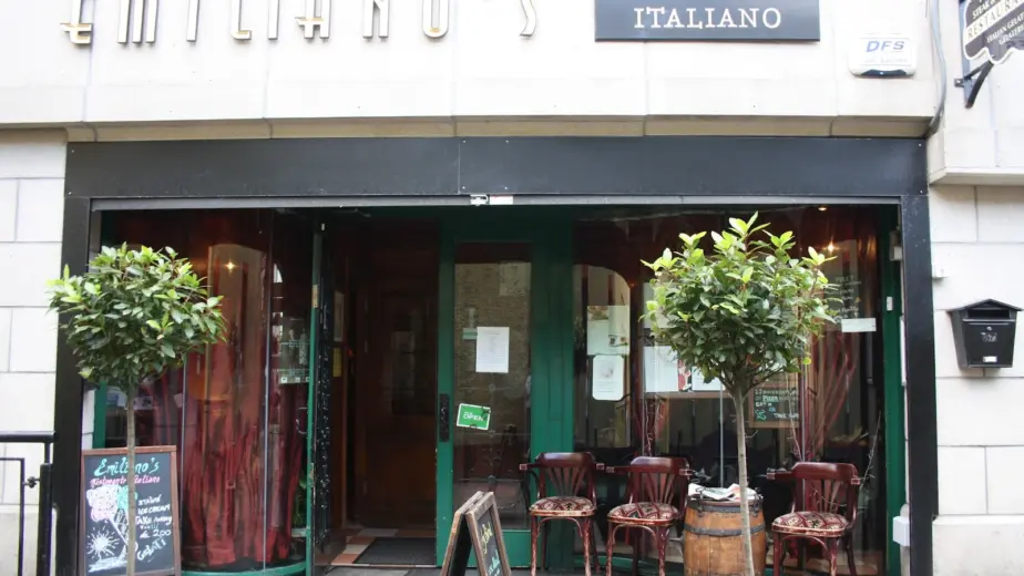 Front entrance of Emiliano’s Ristorante Italiano with glass doors, potted trees, wooden chairs, a wine barrel, and a restaurant sign above—perfect to visit as you explore Waterford St Patrick’s Day Festival nearby.