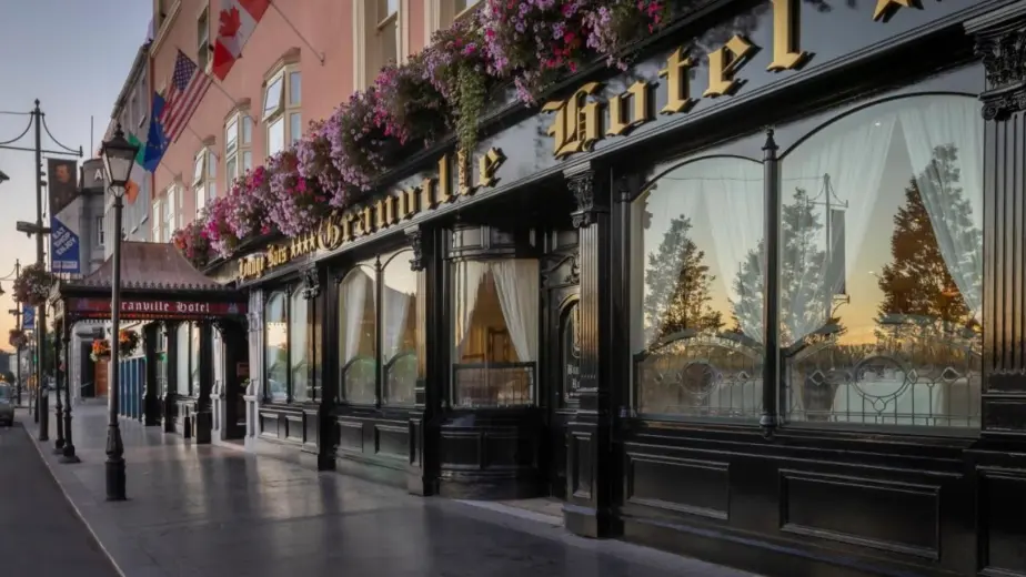 Street view of the Granville Hotel’s ornate black and gold exterior, with flower boxes, flags above, and large windows reflecting trees and sky. The hotel sign and entrance invite you to explore Waterford St Patrick’s Day Festival nearby.