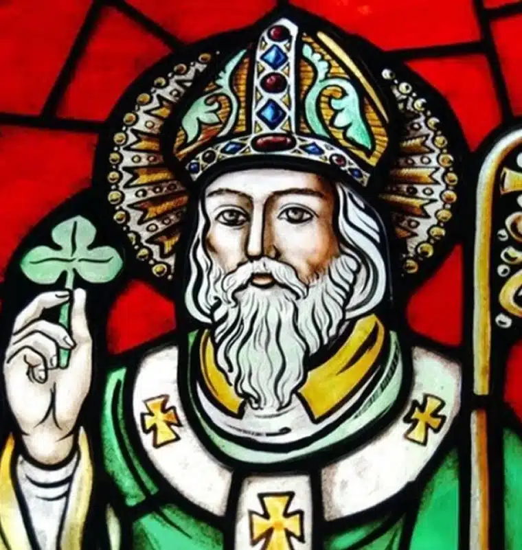 Stained glass depiction of a bearded man in ornate religious robes and hat, holding a shamrock and bishop’s staff, set against a vivid red background—an iconic tribute to the history of St Patrick in Ireland.