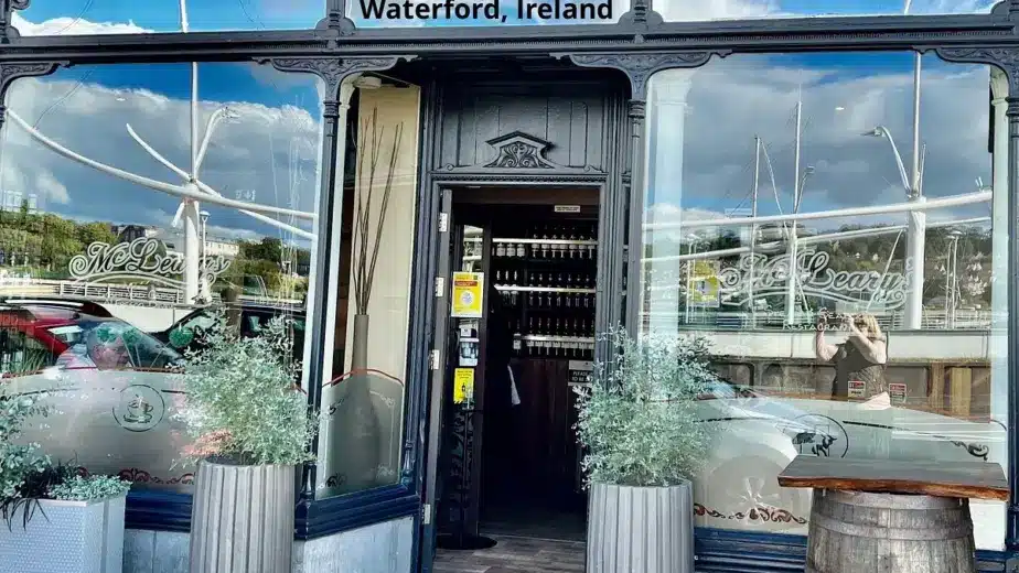 The entrance of McLeary's restaurant in Waterford, Ireland, with large windows, two tall planters, a wooden barrel, and reflections of boats visible in the glass—an inviting spot to explore during the Waterford St Patrick’s Day Festival.