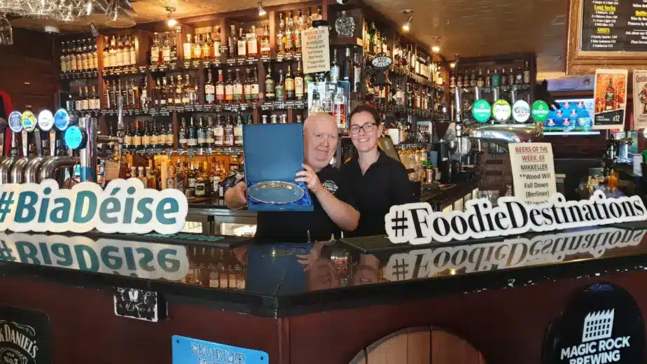 Two people stand behind a bar holding a glass award and smiling. The bar is filled with bottles and taps, and large signs say "#BiaDéise" and "#FoodieDestinations." Discover the charm—Explore Waterford St Patrick’s Day Festival in this warm, inviting setting.