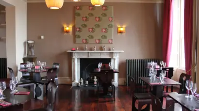 A cosy restaurant dining area with dark wooden tables and chairs, set for service. After you explore Waterford St Patrick’s Day Festival, relax by the white fireplace beneath floral wall art, under modern lights, near windows draped in red curtains.