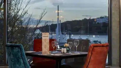 A restaurant table set for four with colourful chairs overlooks a marina with yachts and water at sunset, seen through large windows. As you dine, imagine exciting moments like the Explore Waterford St Patrick’s Day Festival.