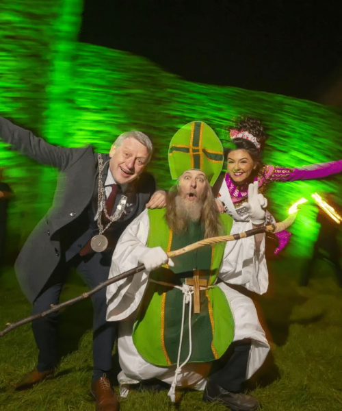 Three people pose playfully outdoors at night, including a man in a suit, someone dressed as Saint Patrick with a long beard and staff, and a young woman in a bright Irish dance costume. Green light illuminates a stone wall behind them.